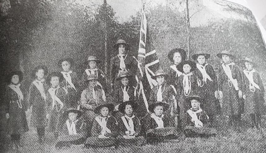A monochrome picture of a Girl Guide Company c1914, wearing white haversacks, broad brimmed hats, navy tunics and skirts, grouped around a Union Flag.