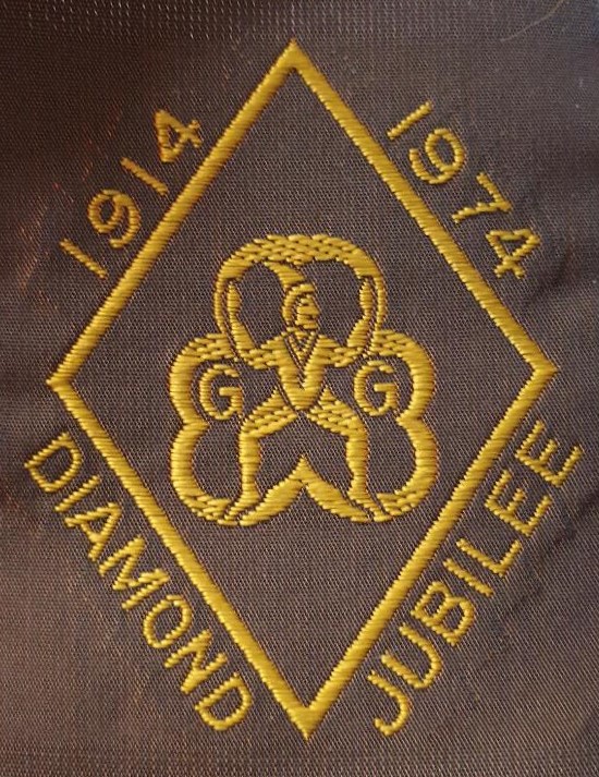 Rectangular brown ribbon badge with gold-stitched Brownie Badge surrounded by a gold diamond, with gold lettering on the sides. Text: 1914 1974 Diamond Jubilee
