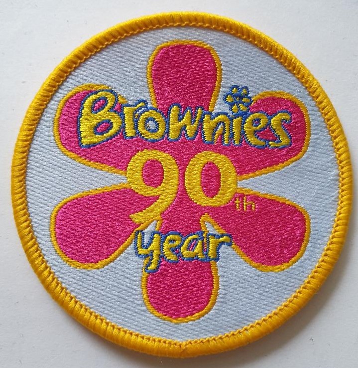 Brownies 90th Year badge featuring a pink 6-petal flower on a light blue background with yellow edging.