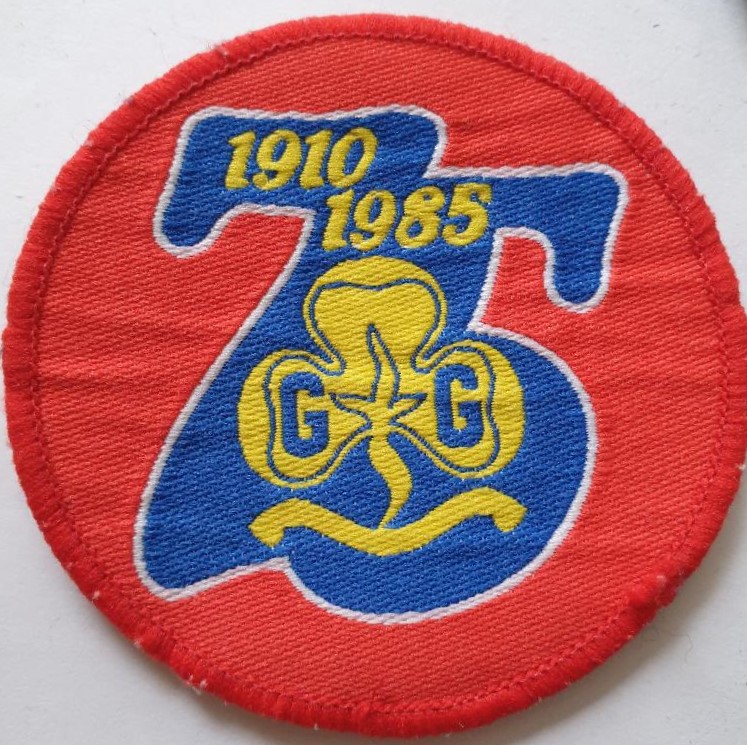 Round red woven badge with red edging, with a central 75-shaped logo in blue piped white. On it a yellow trefoil and text: 1910 1985