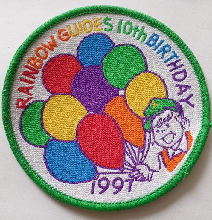 Rainbow 10th Birthday Badge featuring a girl in orange tabard and green baseball cap holding 10 coloured balloons
