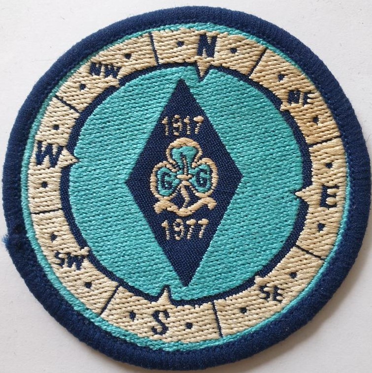 Round cloth badge in aqua with a navy central diamond featuring trefoil in white with aqua infill and white text: 1917 1977. Surrounded by a white ring with compass markings in navy, and navy binding..