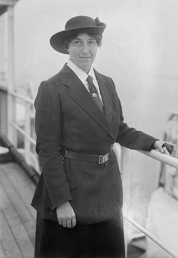 Photo of Olave Baden-Powell in early Guiding Leader uniform with brimmed hat, navy long jacket, and brown leather belt, on the deck of a ship.