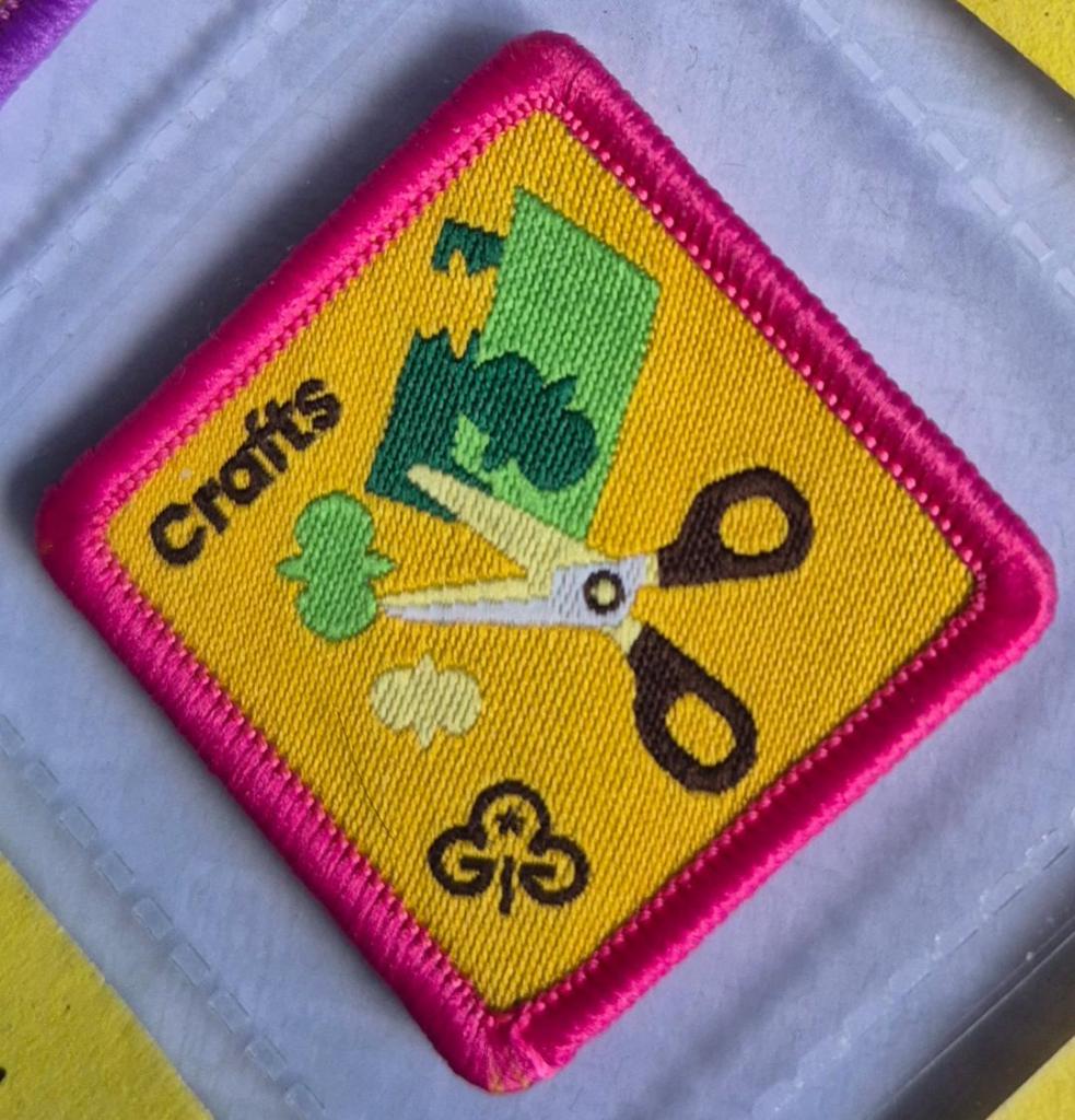 Brownie Crafts badge, dark pink binding, logo shows scissors cutting paper.